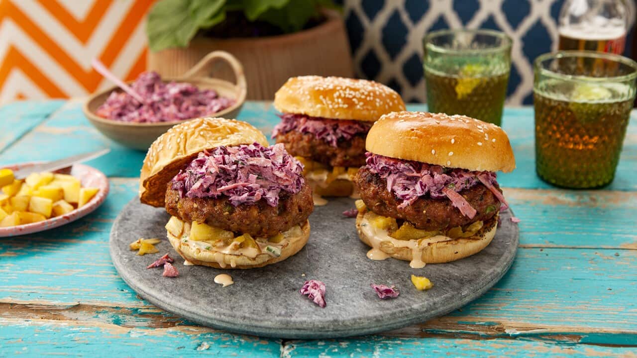 Three burgers sit on a round grey platter, each with a chunky patty and purple slaw. A bowl of slaw sits behind, along with drinks in glasses.