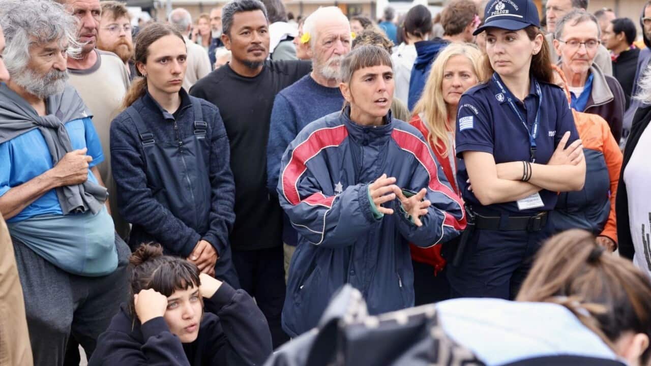 A group of people gather around two police officers in a public outdoor area with a large building and vehicles in the background.