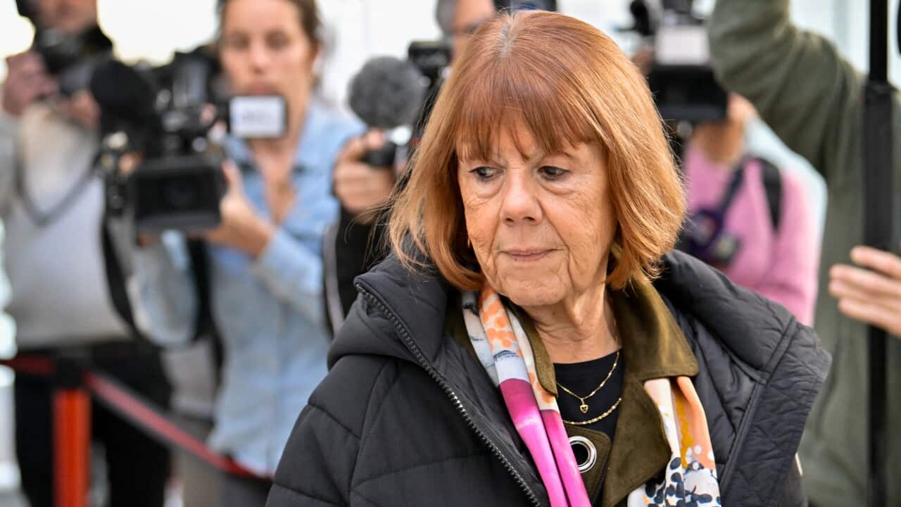 A woman with orange hair wearing a black jacket and a sombre expressions walks in front of several camera operators.