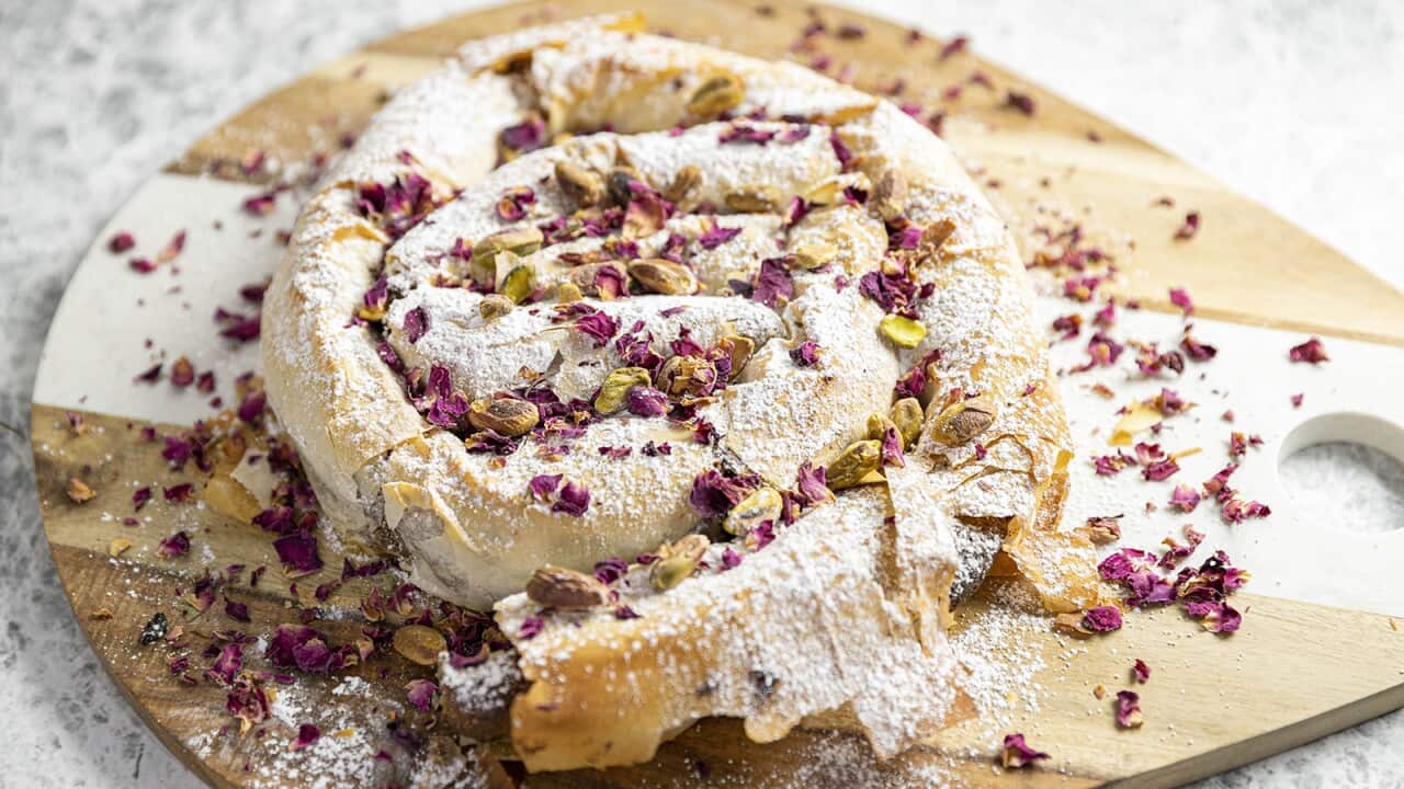 A coiled filo pastry sits on a wooden board. The coil is dusted with icing sugar and decorated with small dried rose petals and pistachios.