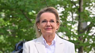 Individual wearing glasses, pearl earrings and a light-coloured blazer stands outdoors before microphones, with leafy trees blurred in the background.