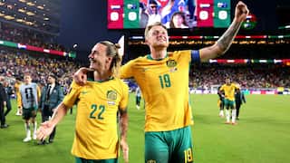 Two Socceroo players embracing each other in celebration with one's hand raised as they are walking across a stadium field.