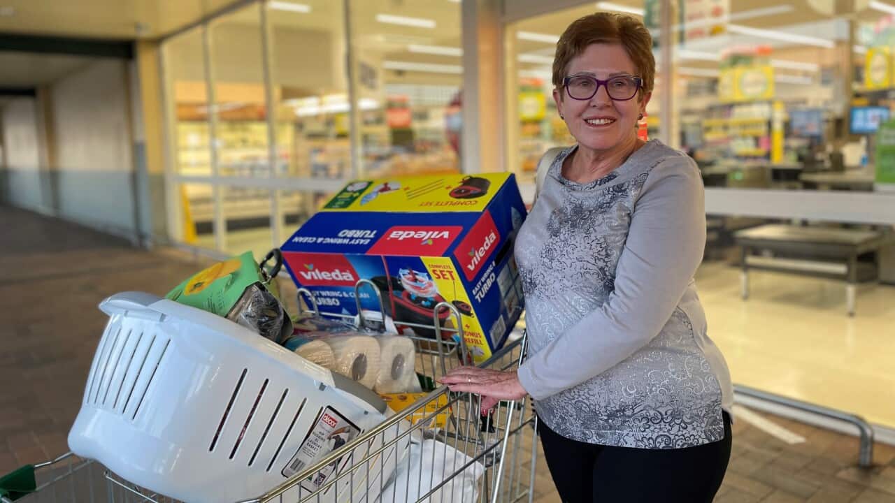 A woman smiles in a grey shirt, touching a trolley full of items in front of Woolworths.