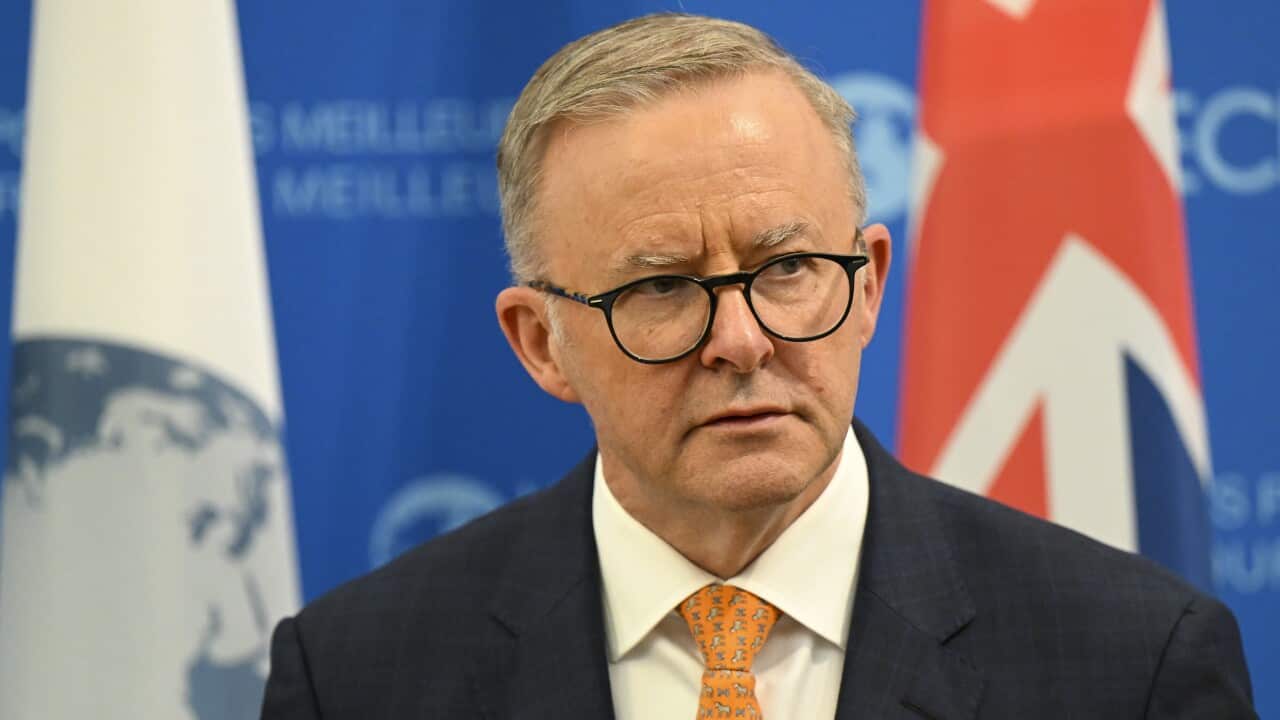 Man wearing a suit and glasses staring away, standing in front of an Australian flag.