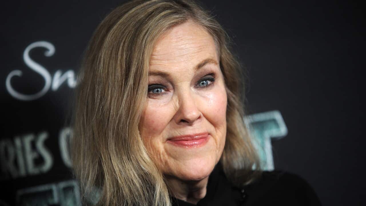A woman with shoulder-length blonde hair smiles softly while posing in front of a dark event backdrop.