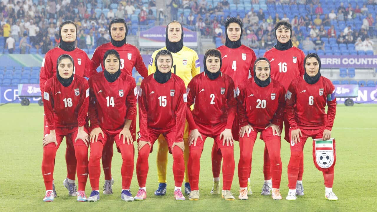 A team of female soccer players from the Iranian national team, wearing red kits and black hijabs, poses for a group photo on a rainy pitch before a match.