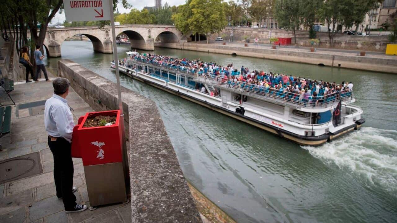 A man stands at a 'uritrottoir' public urinal on August 13, 2018, on the Saint-Louis island in Paris, as a 'bateau mouche' tourist barge cruises past.