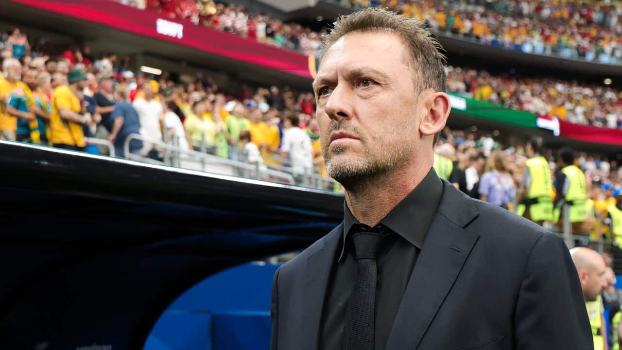 Tony Popovic in a black suit and tie looking concerned in front of a sea of green and gold inside Dallas stadium