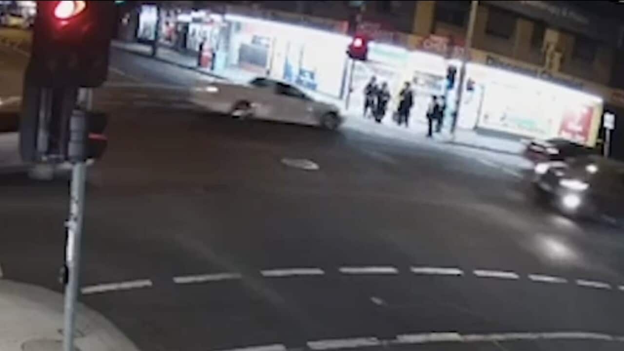 Low-quality CCTV footage captures a white vehicle and a dark SUV navigating a city intersection at night, with several people standing on the sidewalk near a brightly lit storefront in the background.