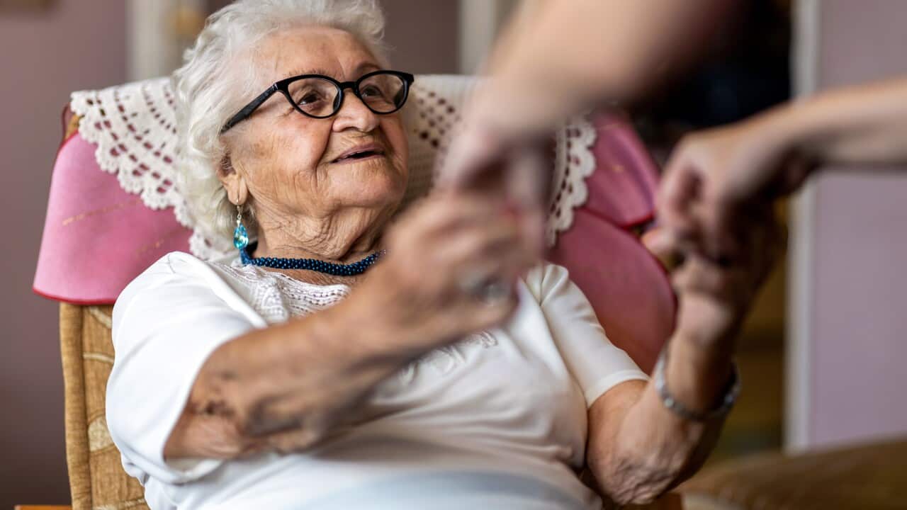 Female home carer supporting old woman to stand up from the armchair at care home