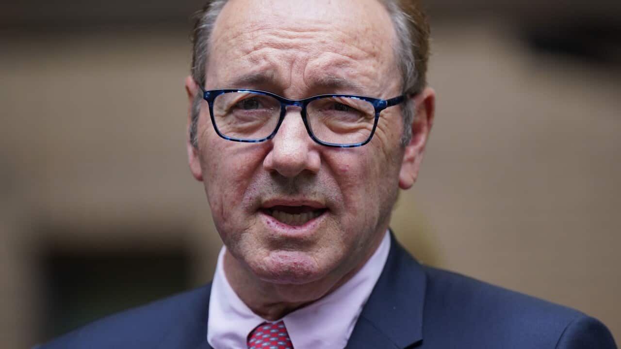 A man wearing a suit, tie and glasses who is speaking.