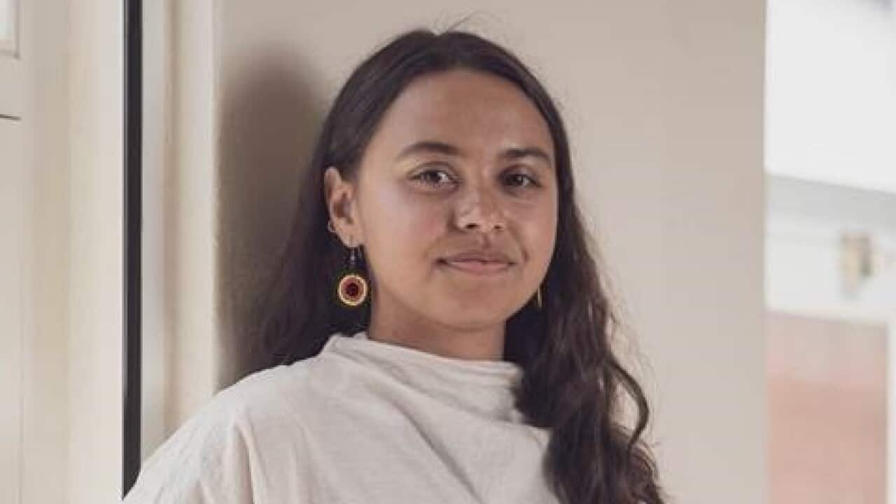 Maya Hodge. She is wearing brown earrings and a beige blouse, in front of a cream wall, looking at the camera.
