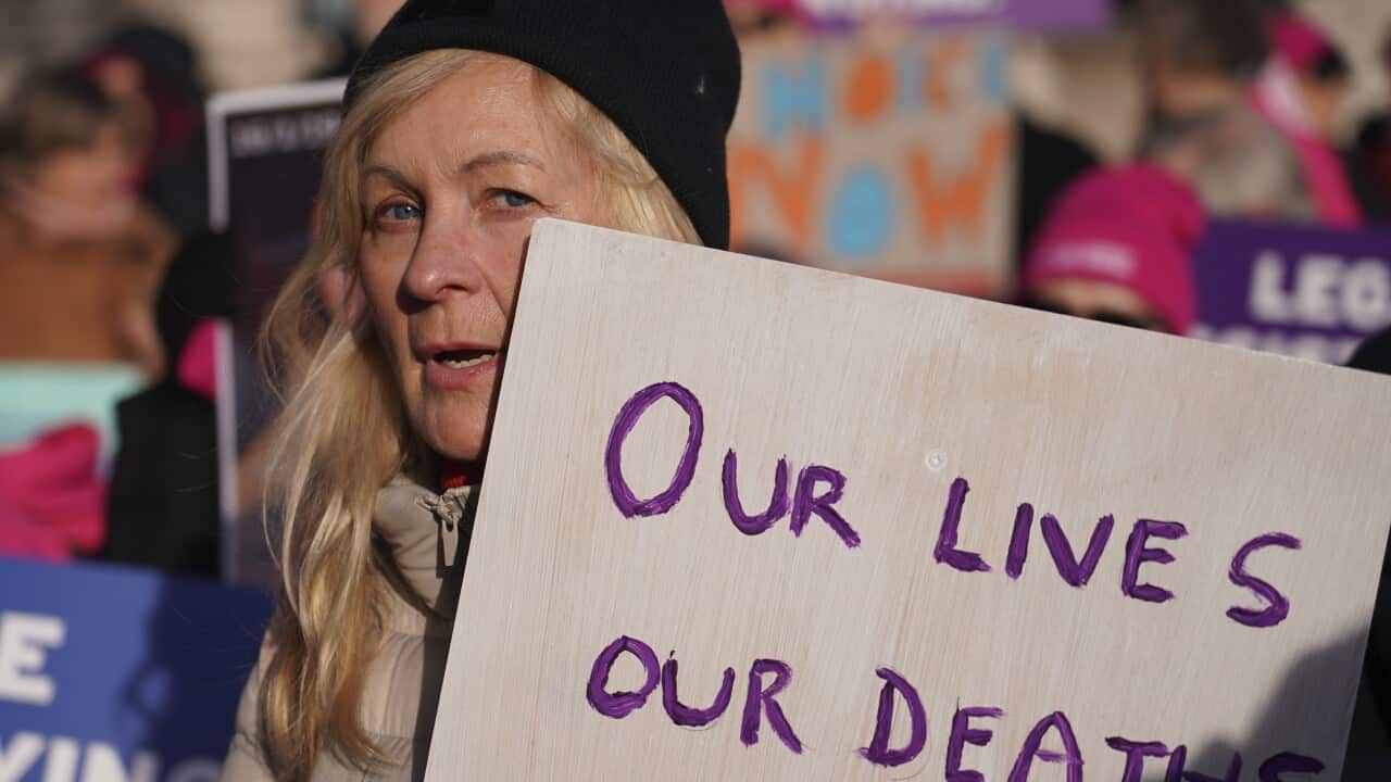 A blonde woman wearing a black beanie holding up a sign that says "Our lives, our deaths".