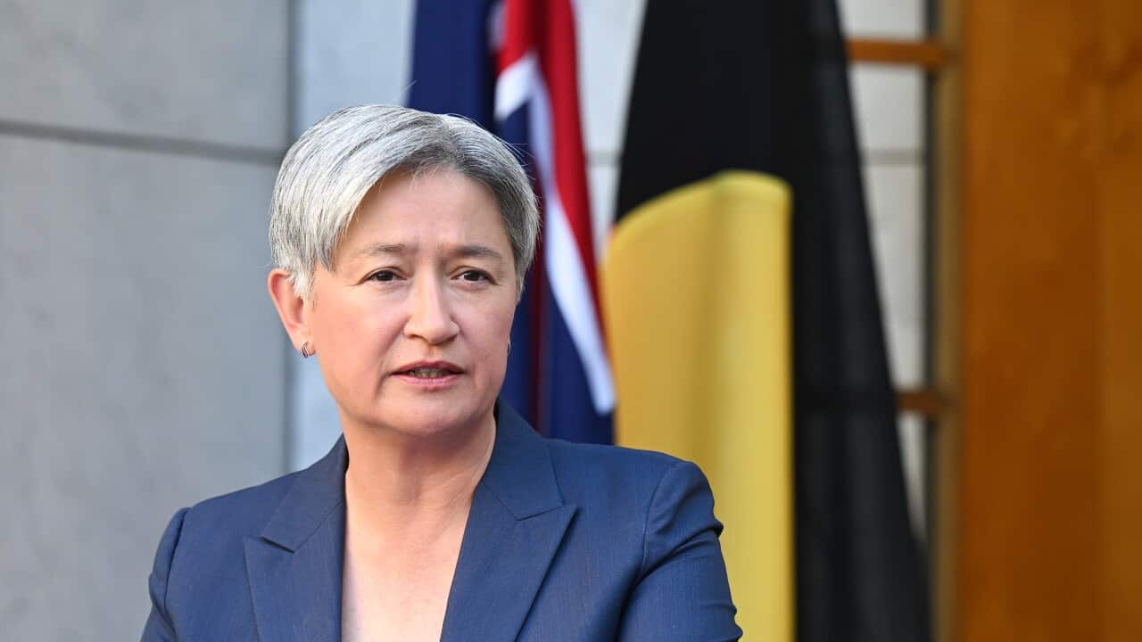Australian Foreign Minister Penny Wong stands in front of the Australian and Aboriginal flags, wearing a blue blazer and black top.