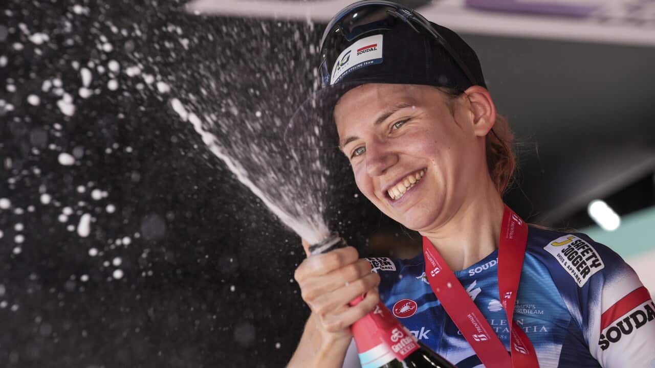 A woman in a blue and white sports jersey and a cycling cap smiles and sprays a bottle of champagne.