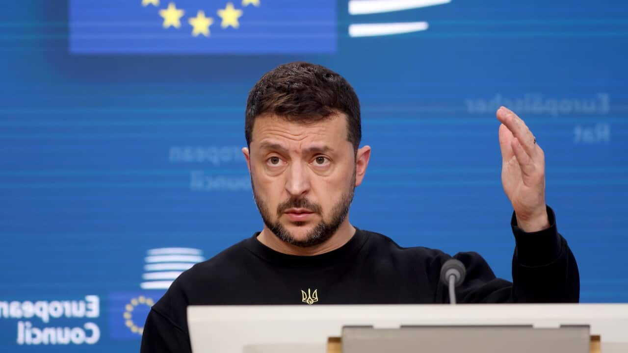 Volodmyr Zelenskyy standing behind lectern. There is a blue background behind him and he is holding up his left hand.