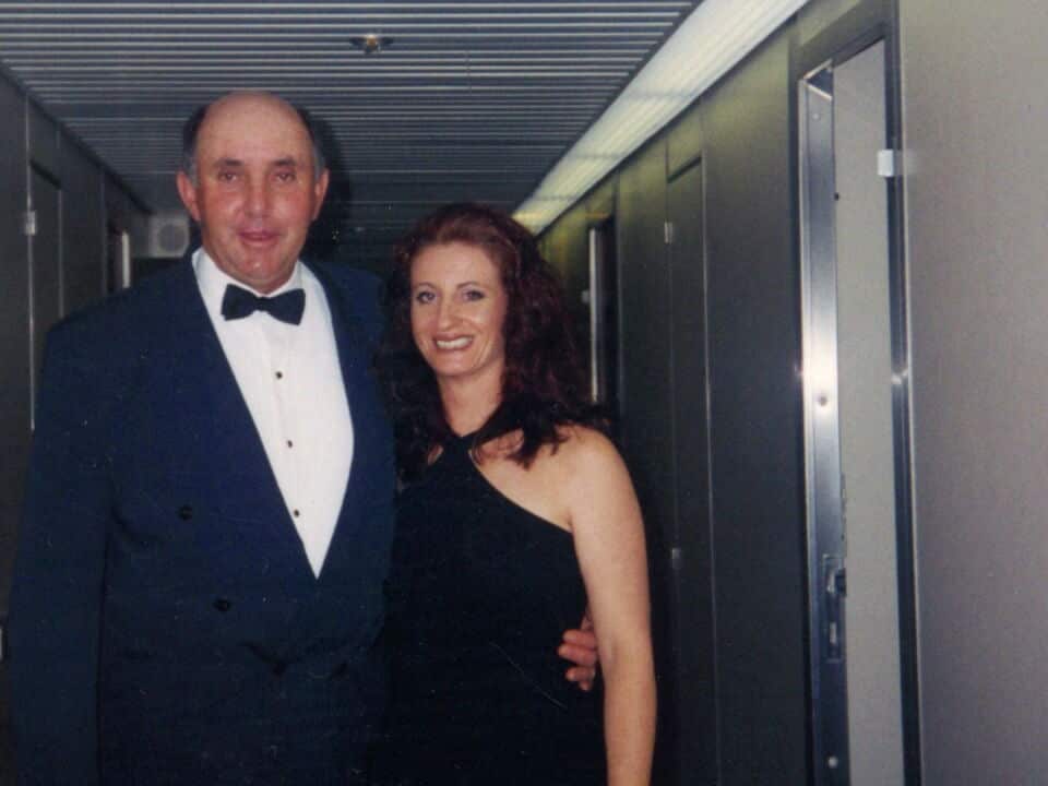 a film photo of a couple in formal wear standing in a dark hallway, the middle aged man has his arm around the younger woman's waist