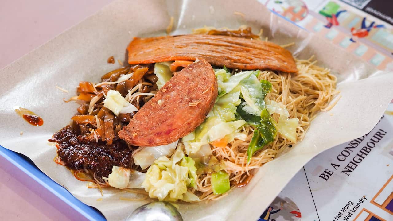 A plate of economy fried beehoon from Singapore, topped with chilli, stir fried cabbage, otak otak and a crispy piece of luncheon meat
