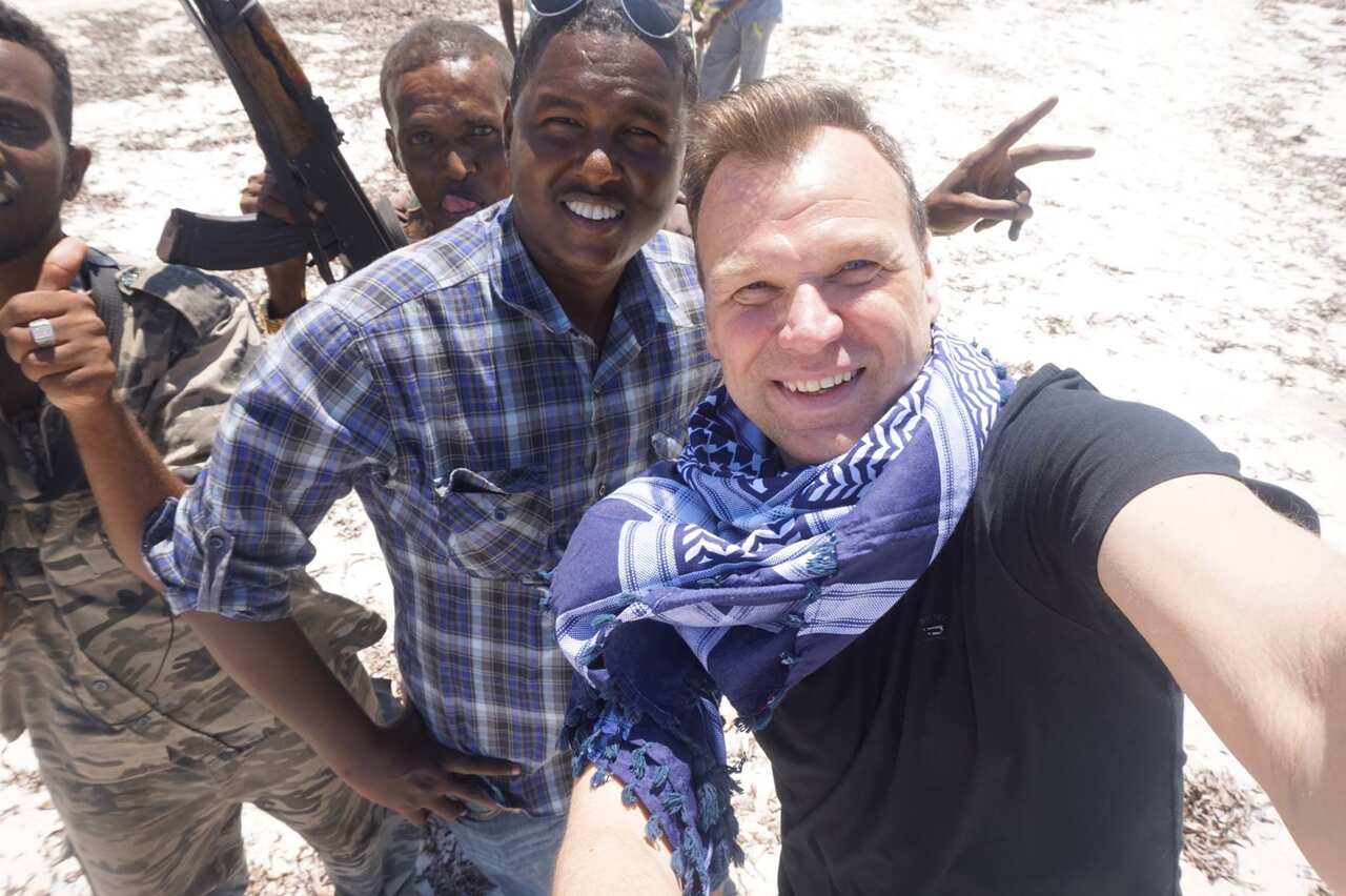 Andrew takes a selfie, a man with a gun poses in the background.