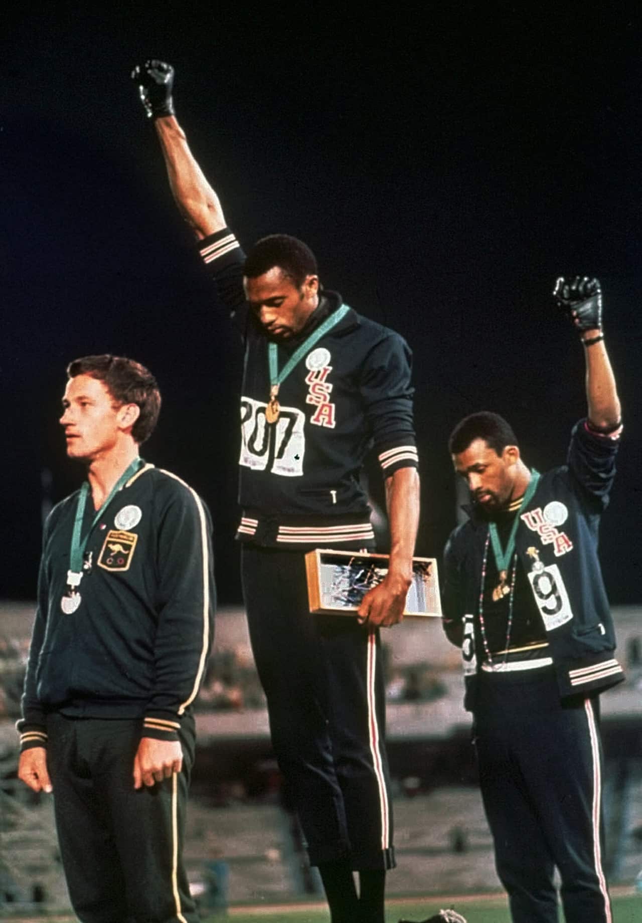 Extending gloved hands skyward in protest, U.S. athletes Tommie Smith, center, and John Carlos stare downward during the playing of the US national anthem.
