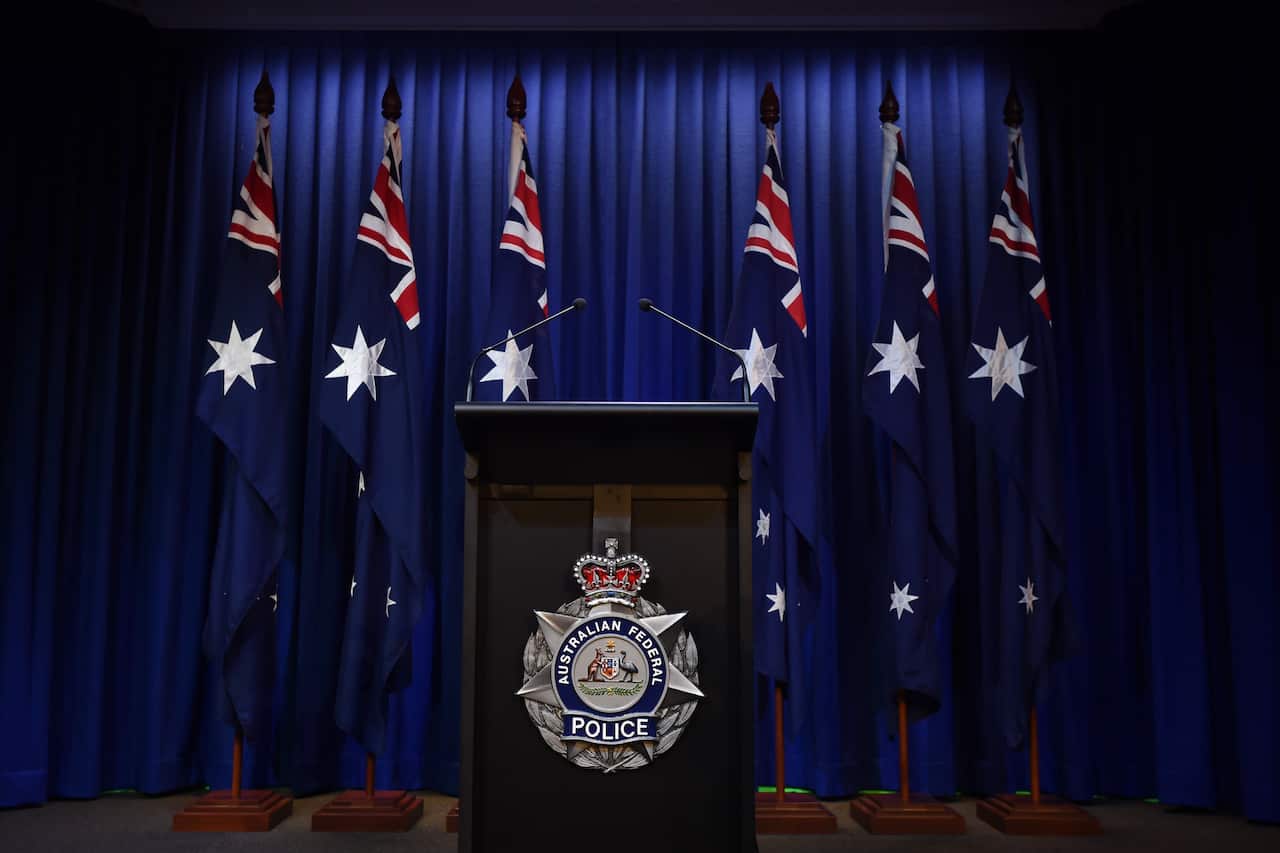 A lectern with the insignia of the Australian Federal Police stands vacant in front of six Australian flags.