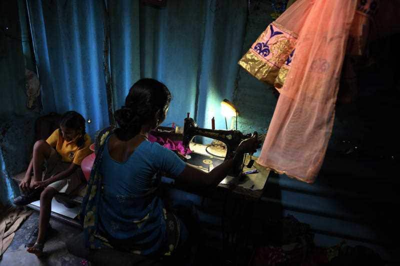 An Indian slum dweller arrives from school as her mother sews clothes under lithium battery run solar lights