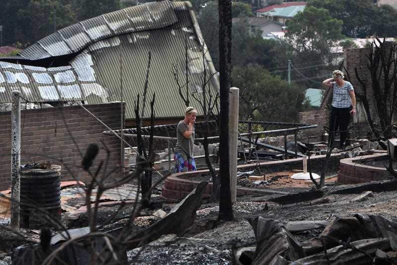 Tathra resident Ingrid Mitchell (left) and Deb Nave (right) wander through the burnt out remains of their Wildlife Drive home in Tathra, Tuesday, March 20, 2018. 