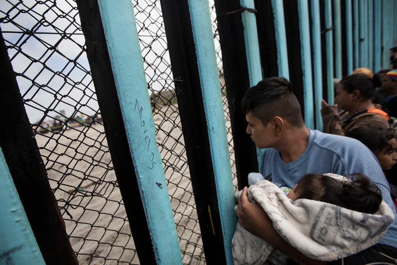 A member of the Central American migrant caravan, holding a child, looks through the border wall