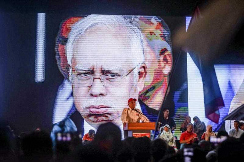 In this May 6, 2018, photo, supporters of former Malaysian strongman Mahathir Mohamad wait as rain pours during an election campaign, with an image of Prime Minister Najib Razak seen on screen.