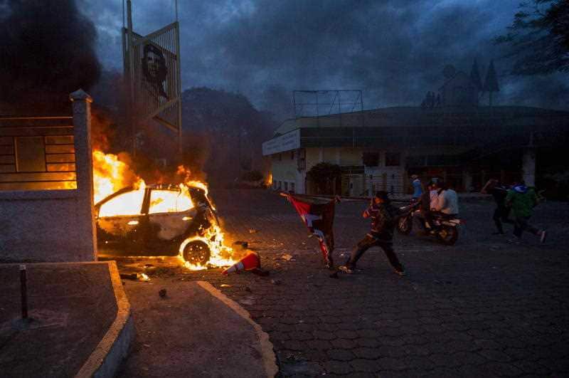 A man throws a flag of the current ruling party towards a burning vehicle as smoke rises from the Caruna Savings and Credit Cooperative headquarters during protests in Managua.