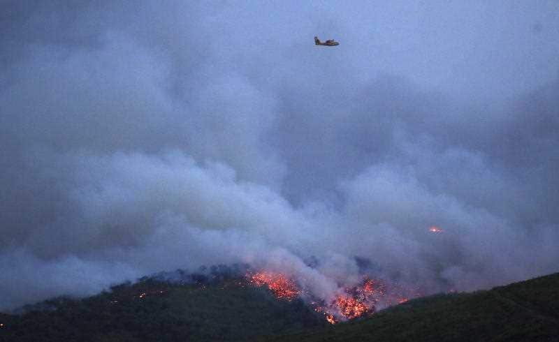 A plane flies over the smoke and fire coming the town of Mati, east of Athens, Monday, July 23, 2018.