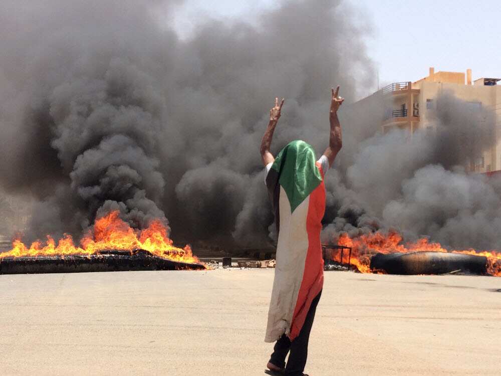 A protester wearing a Sudanese flag in front of burning tires and debris on road 60, near Khartoum's army headquarters