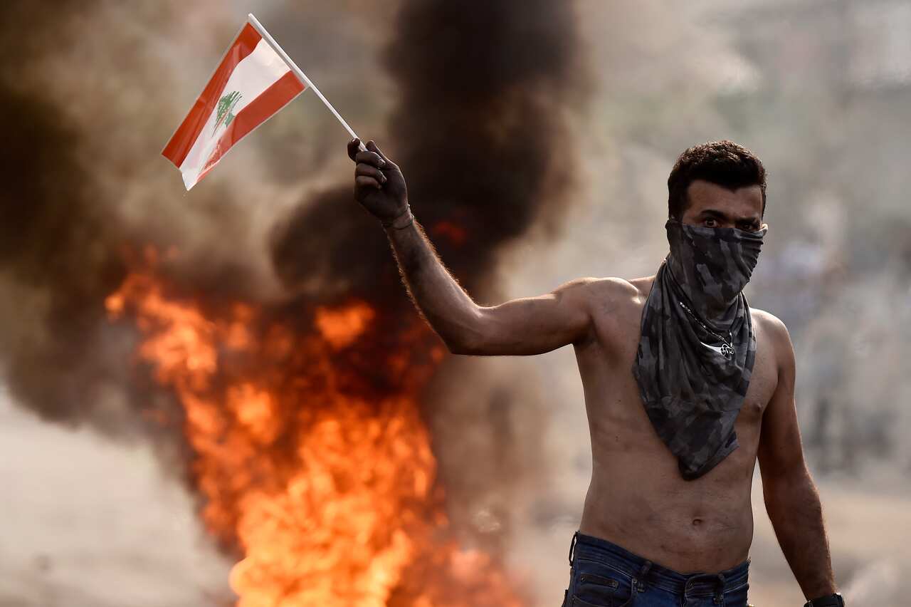 A protester carries a Lebanese flag in front a fire of plastic barriers and trash.