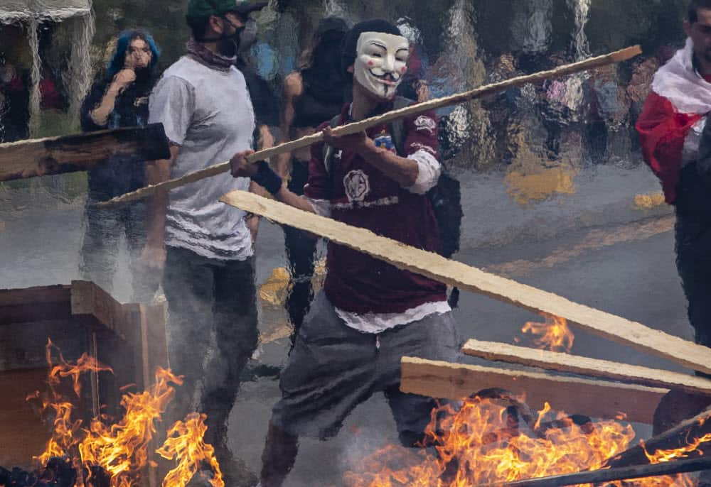 A demonstrator wearing a mask throws a plank of wood into a flaming barricade