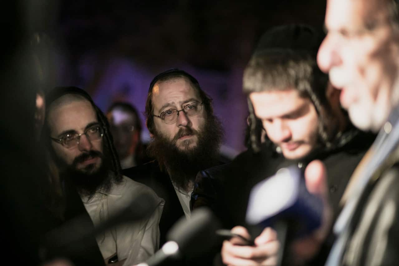 Onlookers, many of them Orthodox Jewish, stand on the street from where five people were stabbed at a Hanukkah celebration.