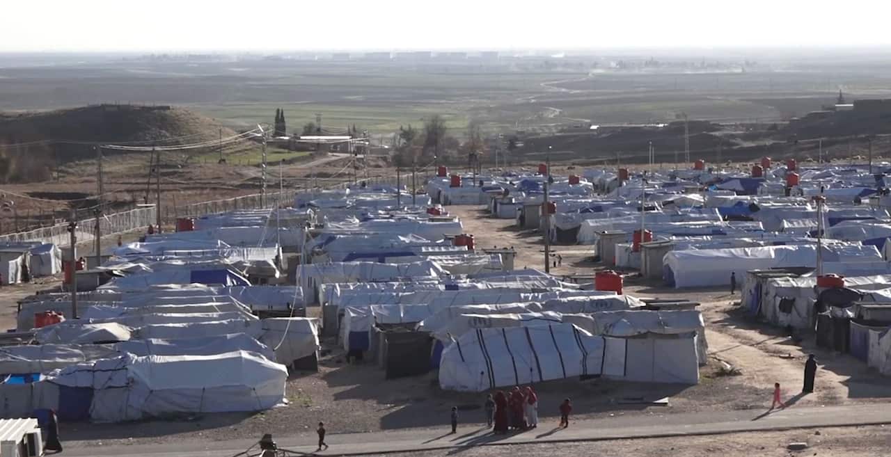 Dozens of white tents stand in the sun within a fenced area surrounded by a dry landscape.