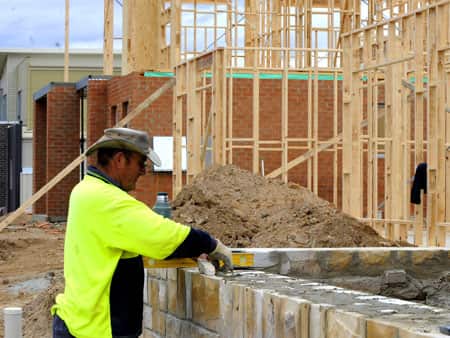 A man in hi vis clothing and a hat is laying bricks at a construction site.