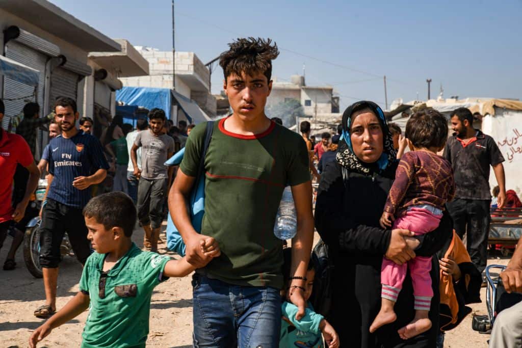 Men, women, and children walk past make-shift brick shelters.