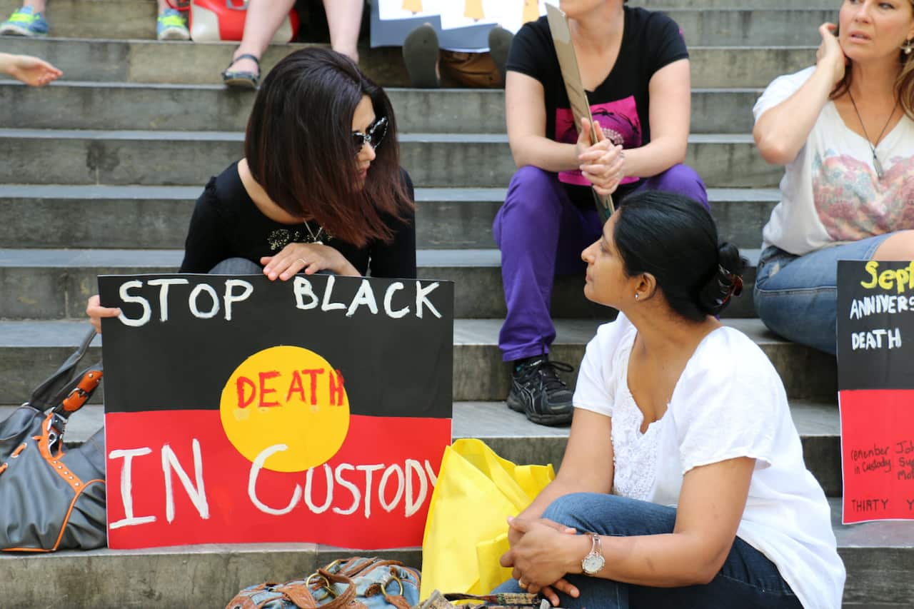 Women sitting on steps. A woman has in front of her a red, yellow and black sign with the words: Stop Black Deaths In Custody written on it in white lettering.