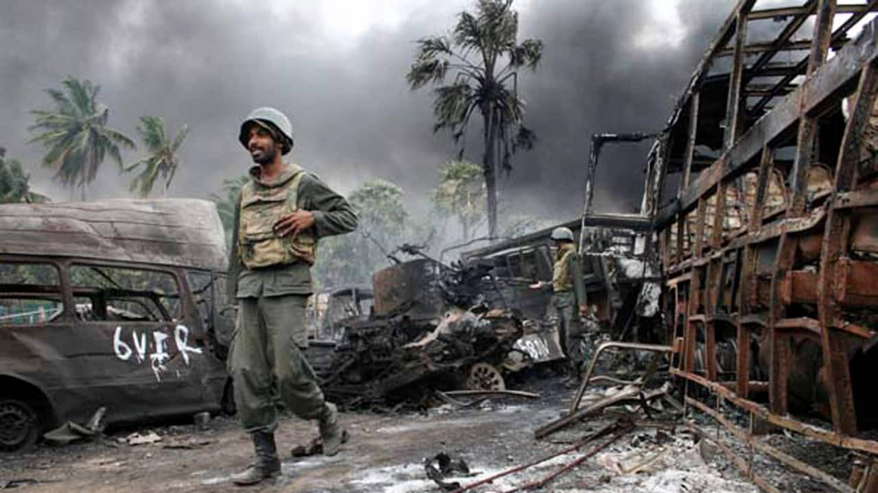 A soldier in green uniform among burnt-out vehicles outside. There is another soldier in a green uniform in the background