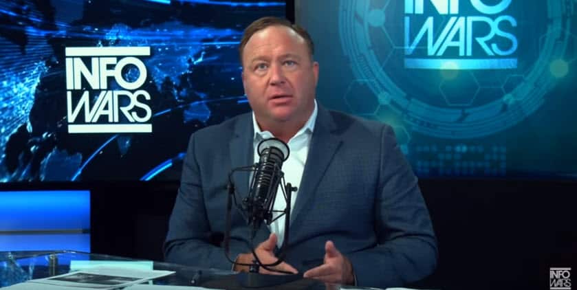 A middle-aged white man on a TV set in front of a microphone. "InfoWars" is on the wall behind him.