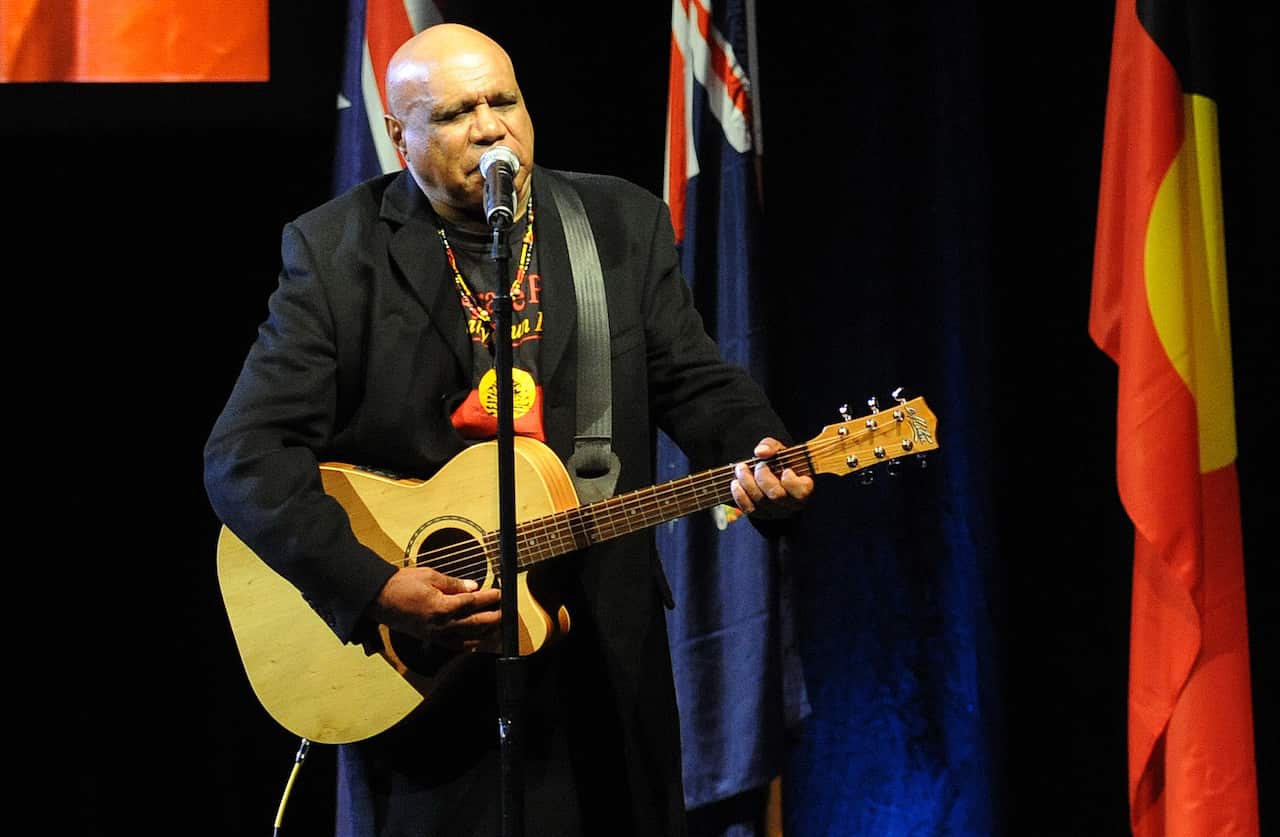 Archie Roach sings on stage, while playing guitar.