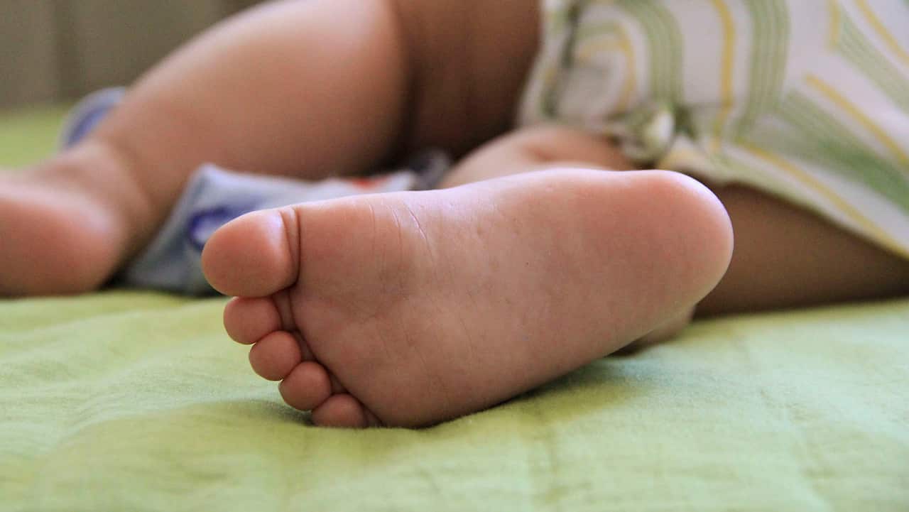 A close-up of a baby's bare foot resting on a bed, with the infant's body softly blurred in the background.