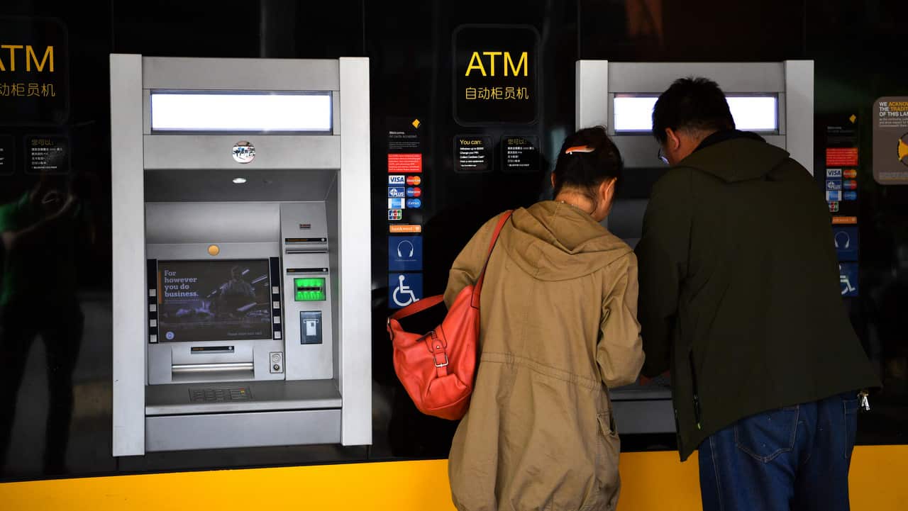 A man and woman wearing a tan jacket stand at a Commonwealth Bank ATM machine.