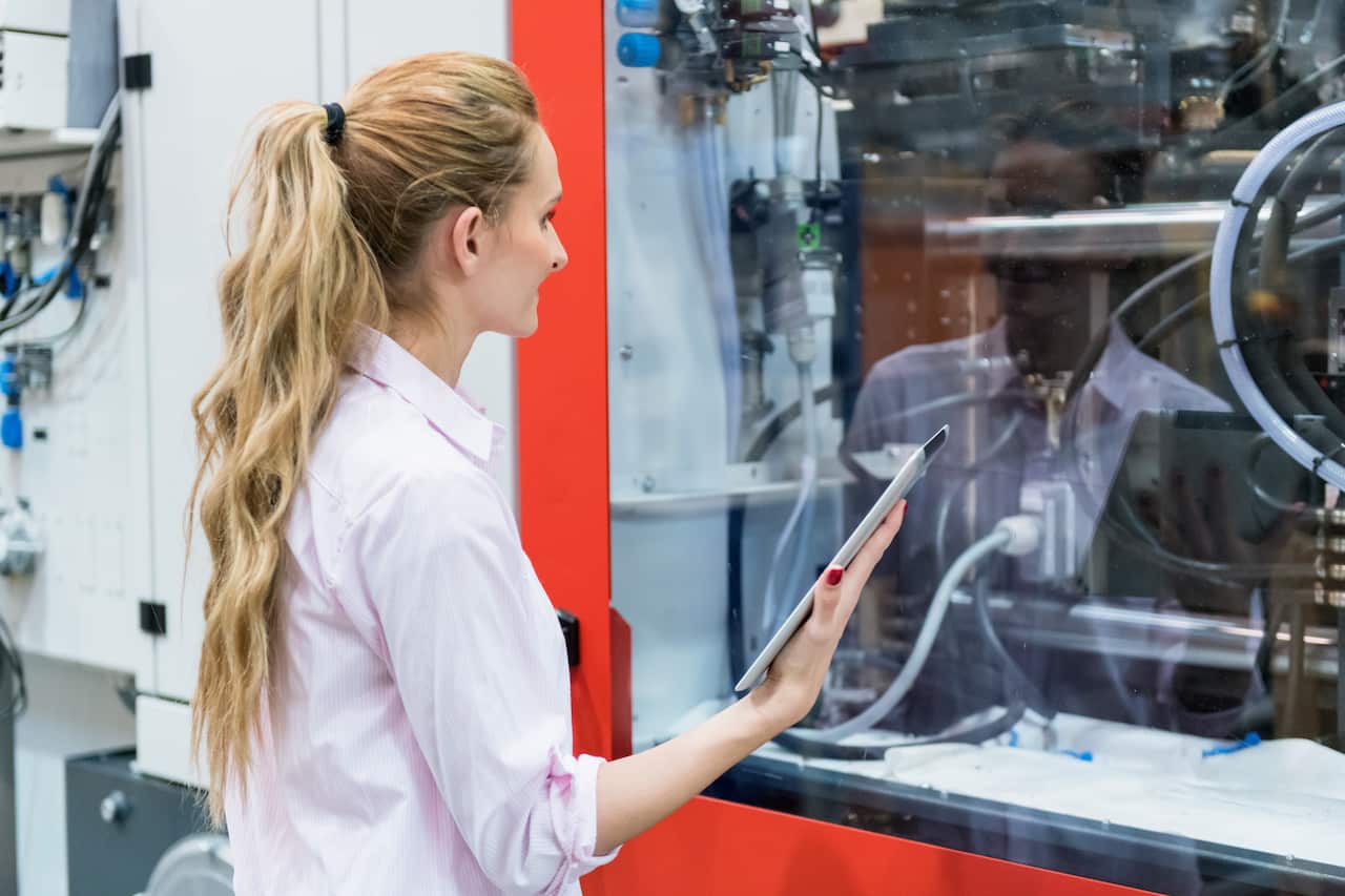 A blonde-haired woman with her hair tied stands in front of equipment inside a glass screen while holding a tablet.