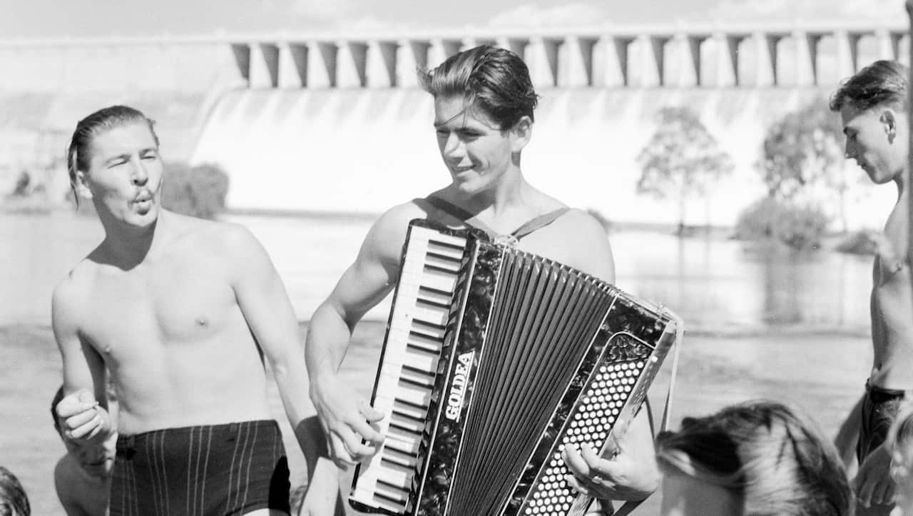 A black and white picture of a man playing a piano accordion outside and a man standing next to him whistling. They are both wearing swimsuits.