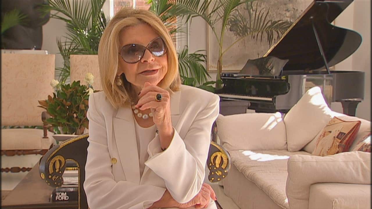 Carla Zampatti wearing a cream jacket and large sunglasses and sitting inside.