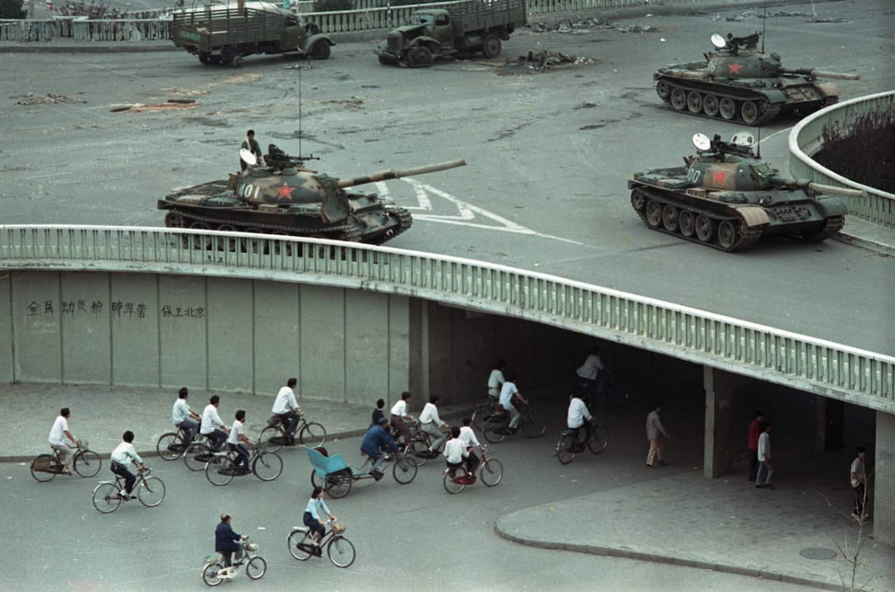 In this June 6, 1989 photo, bicycle commuters, sparse in numbers, pass through a tunnel as above on the overpass military tanks are positioned in Beijing (AAP)