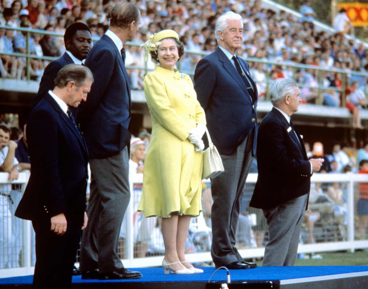 Queen Elizabeth II standing on a stage wearing a yellow outfit and hat and looking at Prince Philip during the closing ceremony of the 1982 Commonwealth Games in Brisbane.