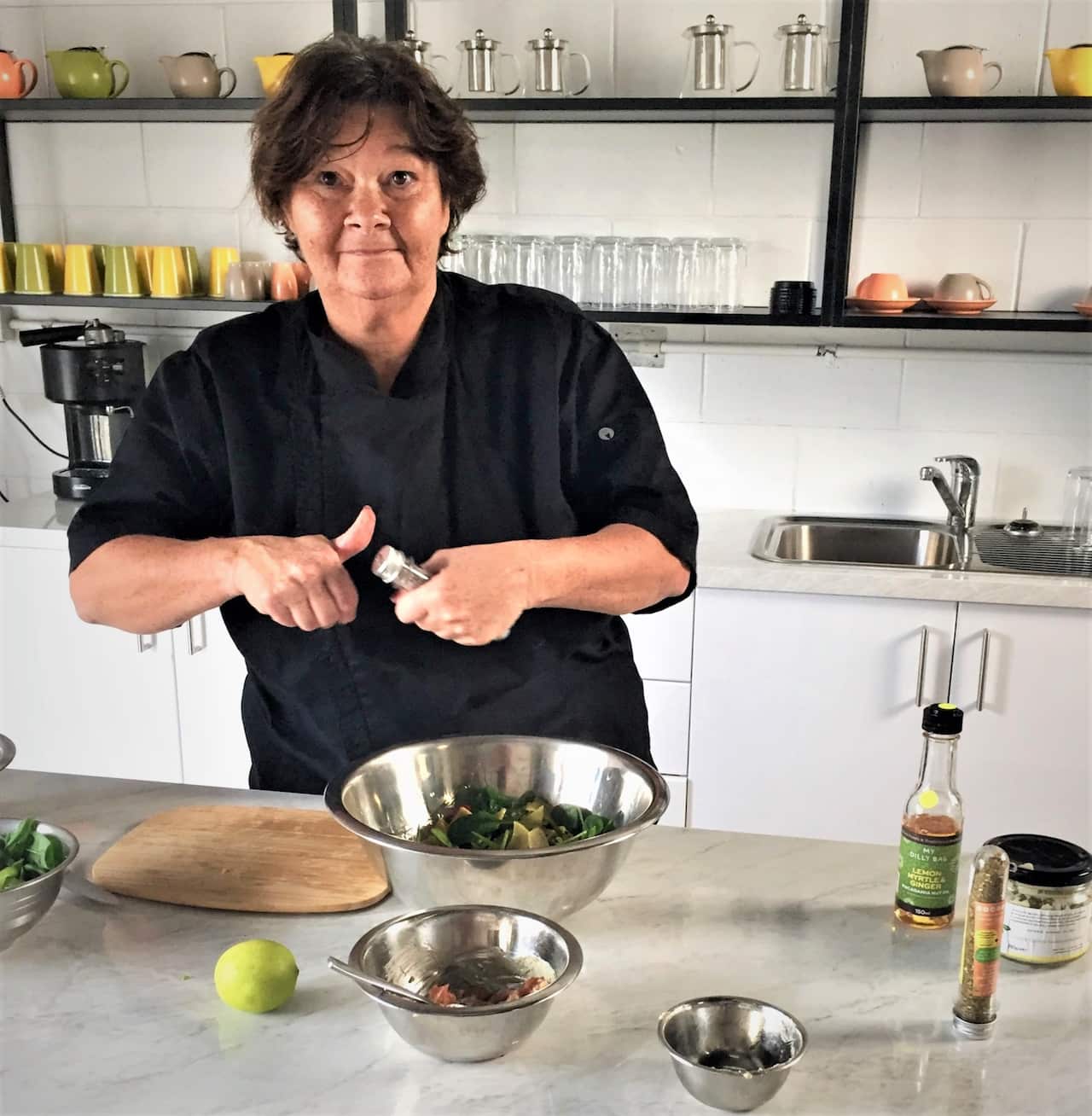 A woman in a black outfit holds a small seasoning shaker jar while a salad bowl is placed on the kitchen island in front of her, and she looks into the camera.